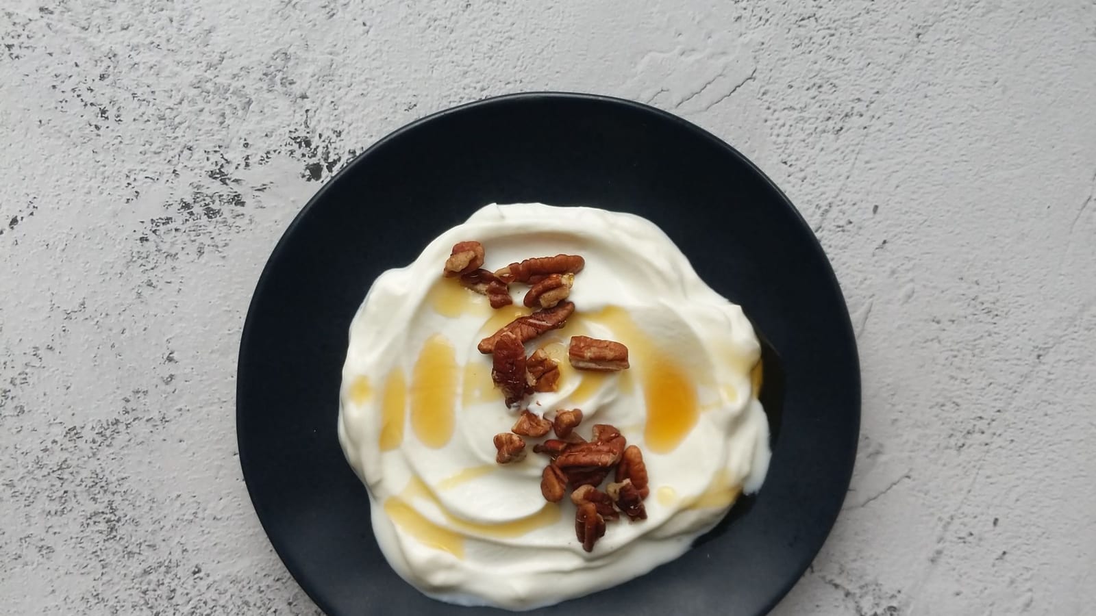 A swirl of thick Greek yogurt topped with honey and chopped pecans on a dark plate.