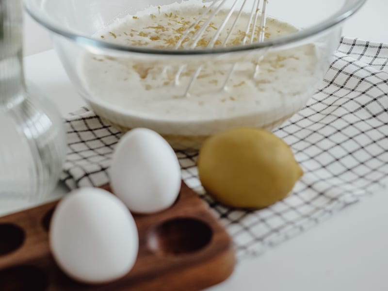 A whisk resting in a bowl of pale whisked egg mixture flecked with lemon zest, a whole lemon and eggs alongside.