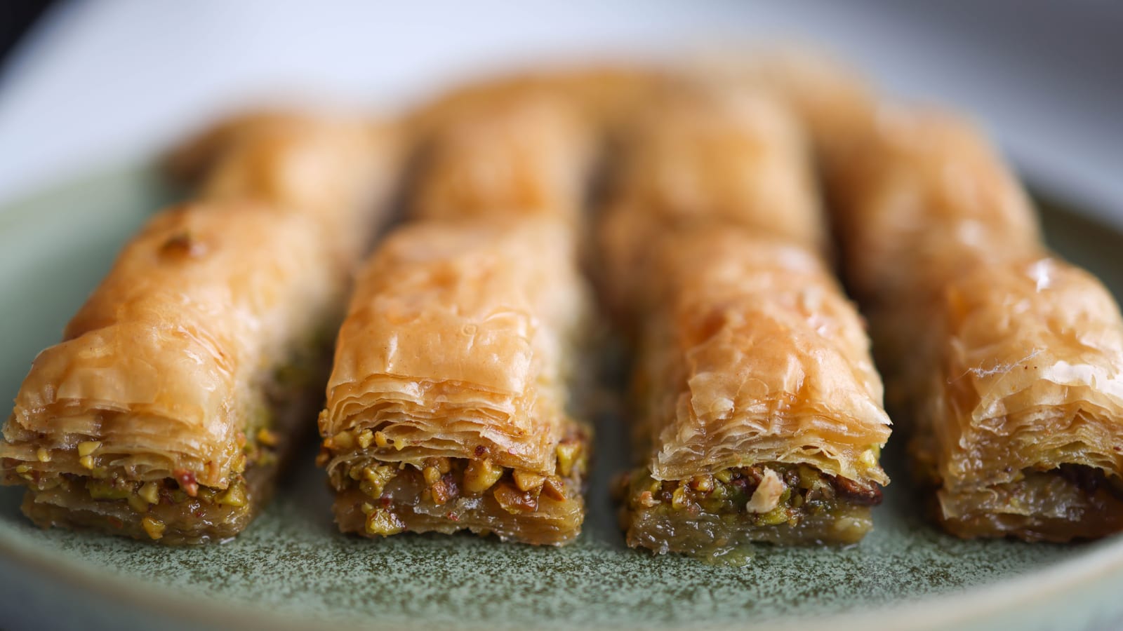 Sliced rolled baklava pieces with pistachio filling on a sage-green ceramic plate, phyllo layers and honey syrup visible.