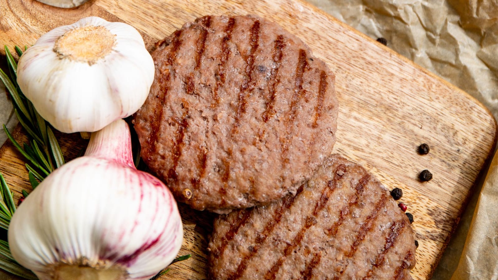 Two grilled beef patties with char marks on a wooden board with cherry tomatoes, garlic, and rosemary.