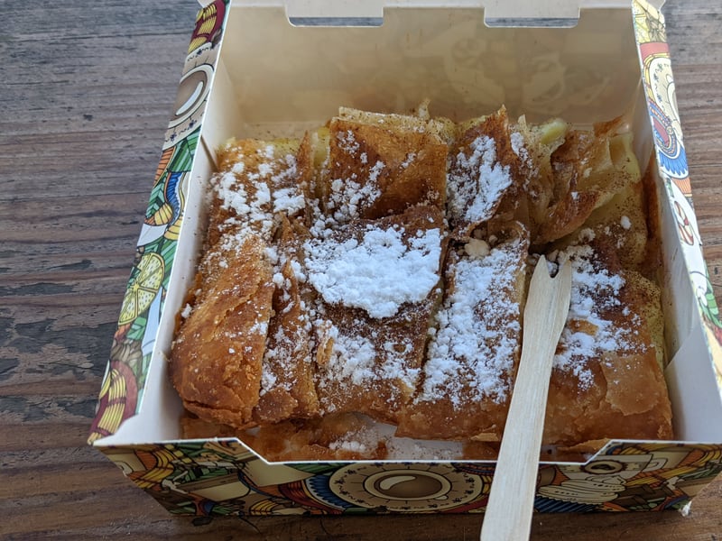 Sliced bougatsa in a bakery takeaway box, dusted with powdered sugar and cinnamon with a wooden fork.