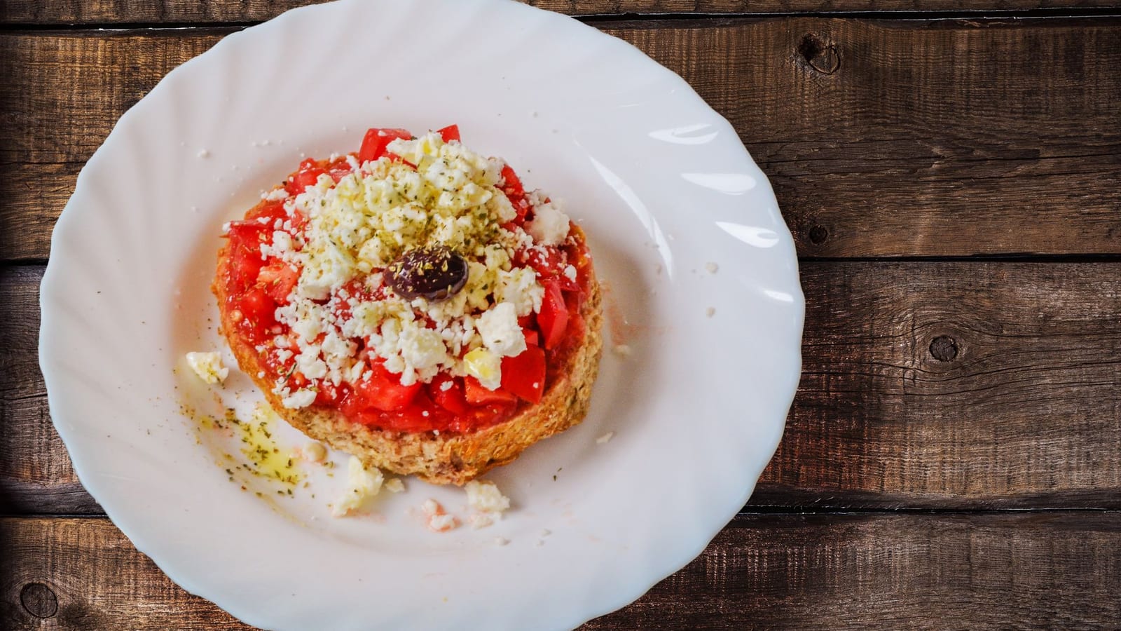 A barley rusk topped with grated tomato, crumbled feta, oregano, and an olive, on a wooden table.