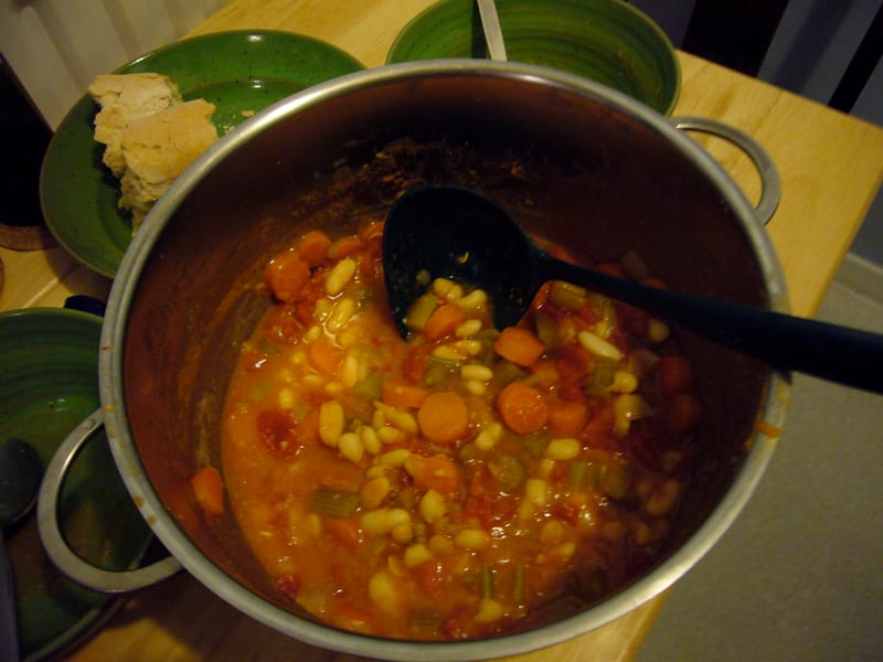 A pot of fasolada simmering on the stove with a wooden spoon resting among the beans, carrots, and celery.