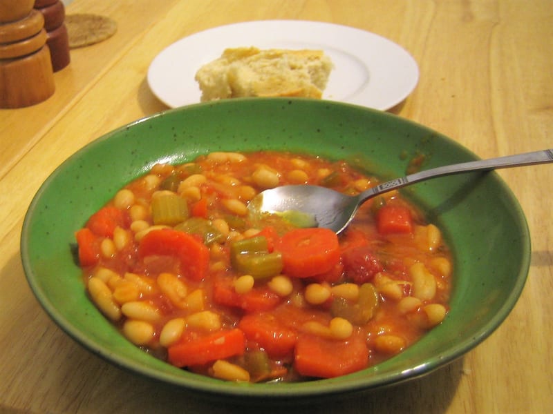 A bowl of fasolada served at the table with a plate of crusty bread and pepper grinders alongside.