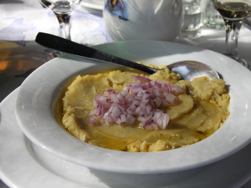 A bowl of fava topped with diced red onion, olive oil pooling around the edges, at a Santorini taverna.