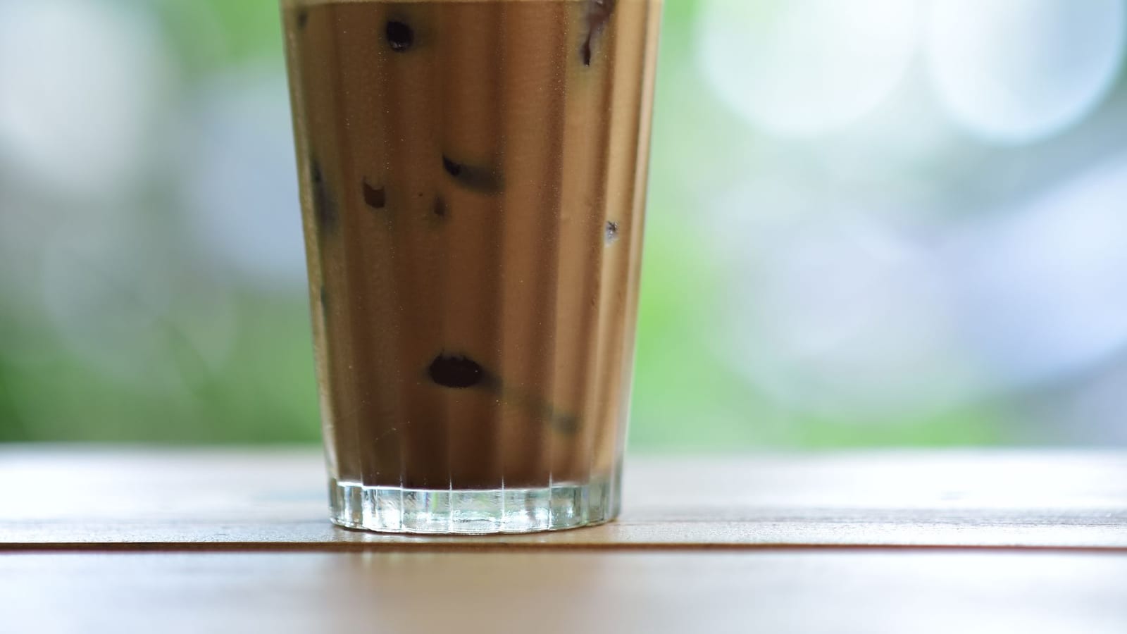 A tall glass of frothy iced coffee with a straw, outdoor greenery blurred in the background.