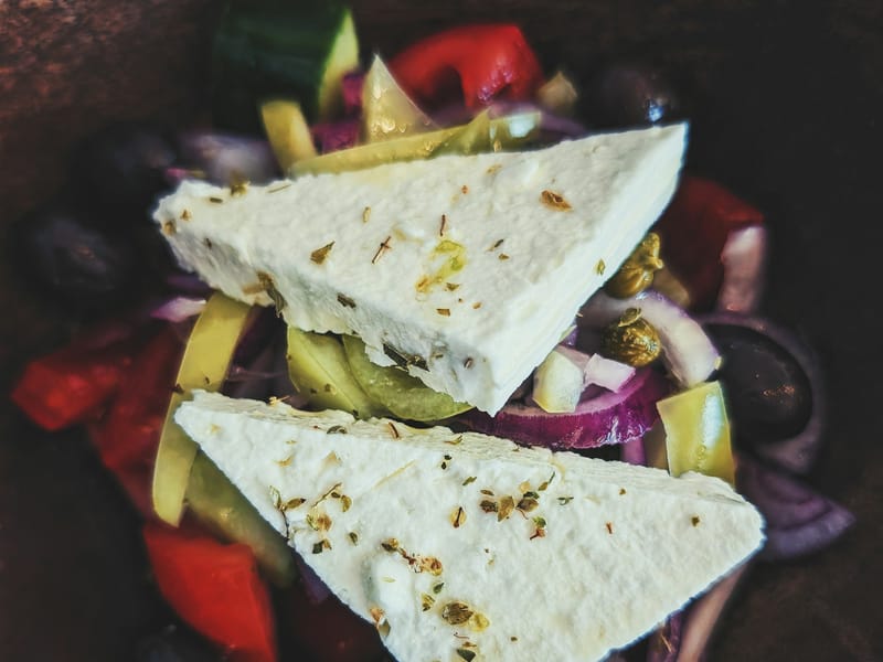 Two thick slices of feta cheese sprinkled with oregano resting on a bed of Greek salad vegetables.