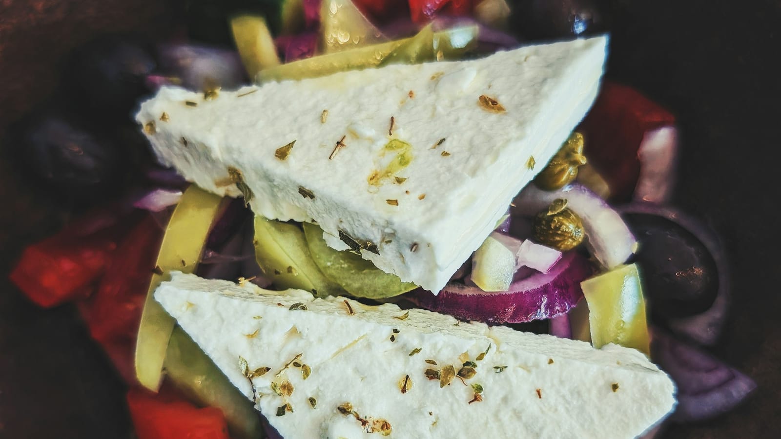 Two thick slices of feta cheese sprinkled with oregano resting on a bed of Greek salad vegetables.