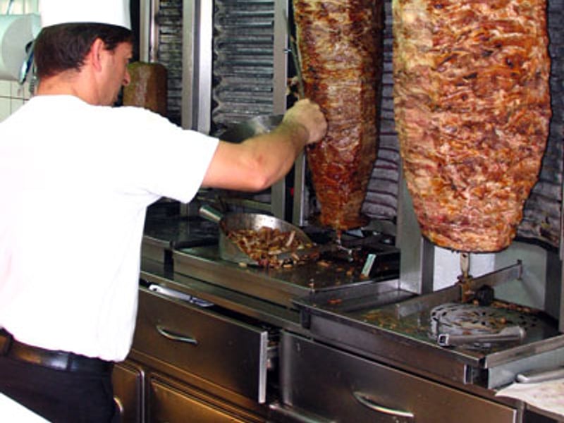 A cook slicing meat off a vertical rotisserie cone in a Greek souvlaki shop.