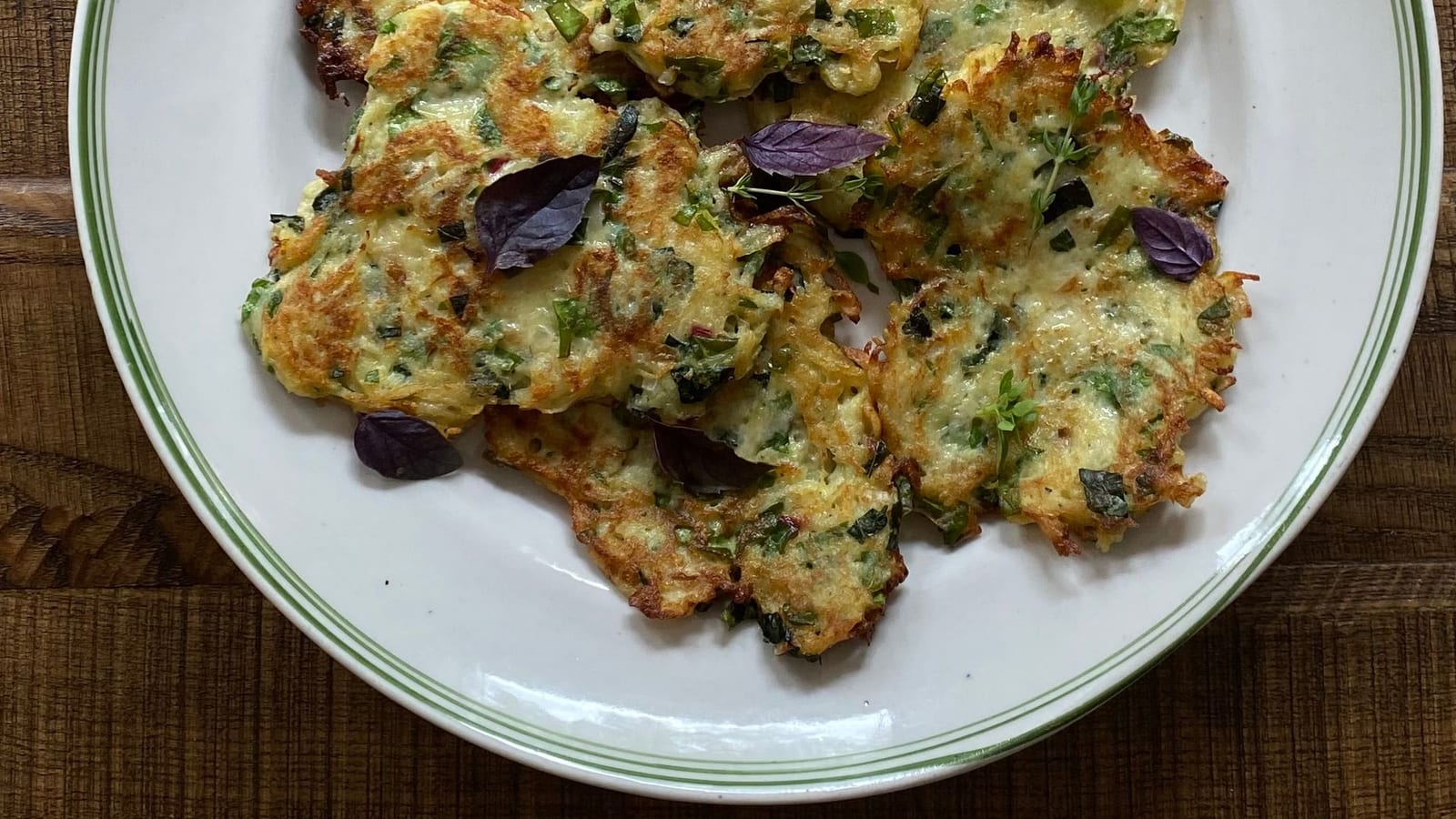 Golden fried zucchini fritters stacked on a plate with fresh herb garnish, on a wooden table.