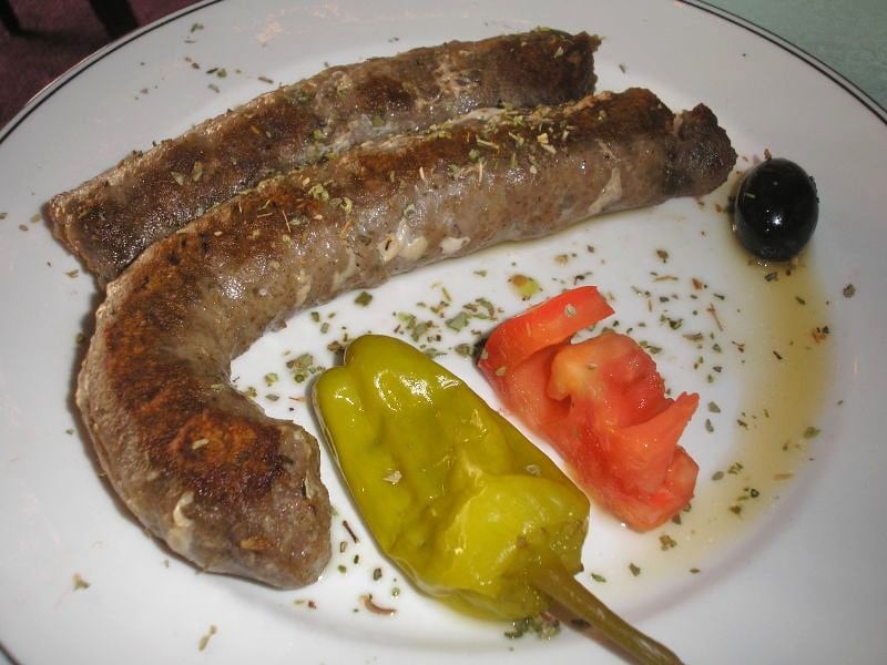 A plate of grilled loukaniko sausage with a roasted pepper, tomato, and an olive.
