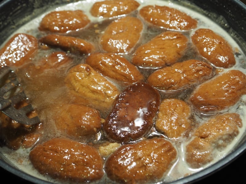 Melomakarona cookies soaking in warm honey syrup in a pan, tongs resting alongside.