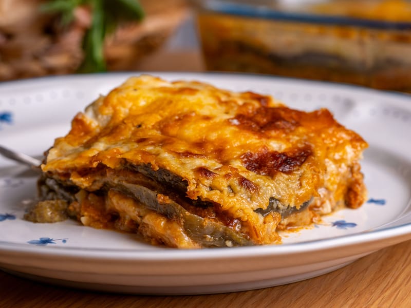 Slice of Greek moussaka with layered eggplant and a golden baked topping, served on a plate with the casserole dish blurred in the background.