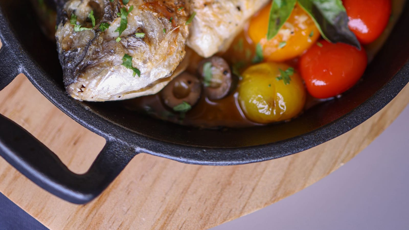 A whole baked fish with cherry tomatoes, olives, and a lemon wedge in a rustic cast iron pot.