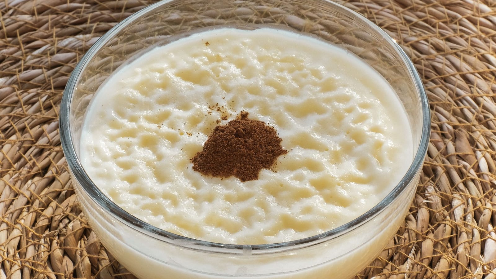 Creamy rice pudding dusted with cinnamon in a glass bowl on a woven mat.