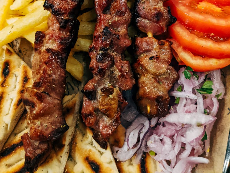 Three grilled souvlaki skewers with charred pork, served over pita with fries, tomato, and red onion.