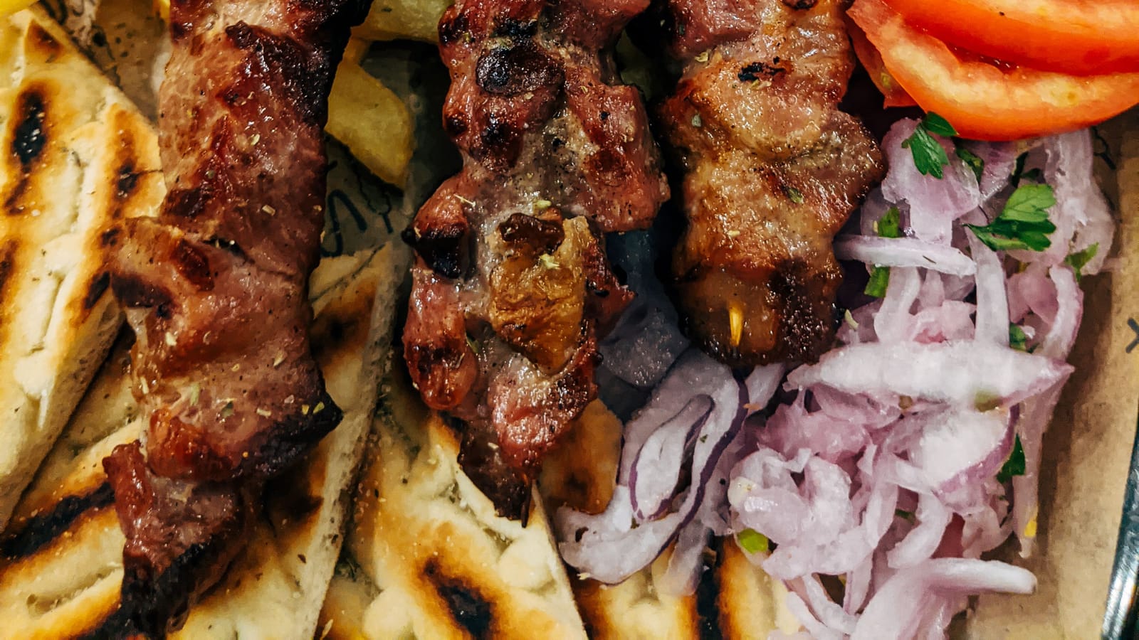 Three grilled souvlaki skewers with charred pork, served over pita with fries, tomato, and red onion.