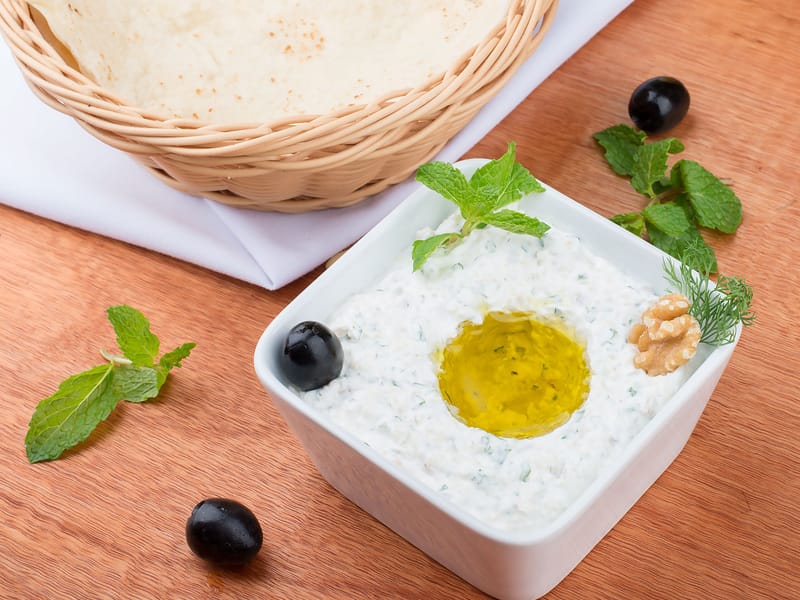 Bowl of tzatziki topped with olive oil, mint, olives, and a walnut, served with a basket of pita bread.